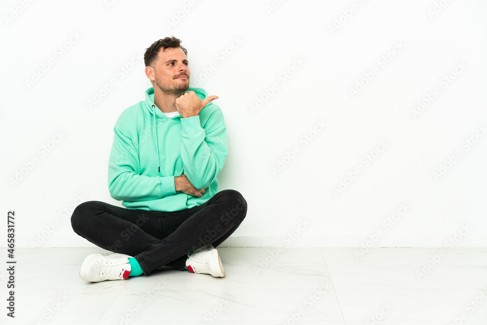 Young handsome caucasian man sitting on the floor pointing to the side to present a product