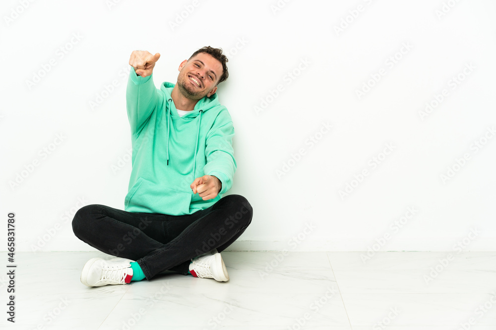 Obraz premium Young handsome caucasian man sitting on the floor points finger at you while smiling