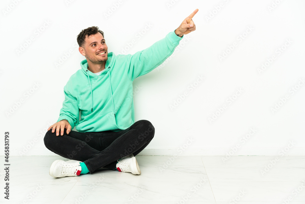 Young handsome caucasian man sitting on the floor pointing away