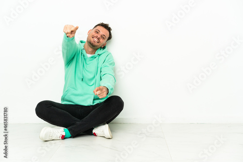 Young handsome caucasian man sitting on the floor points finger at you while smiling