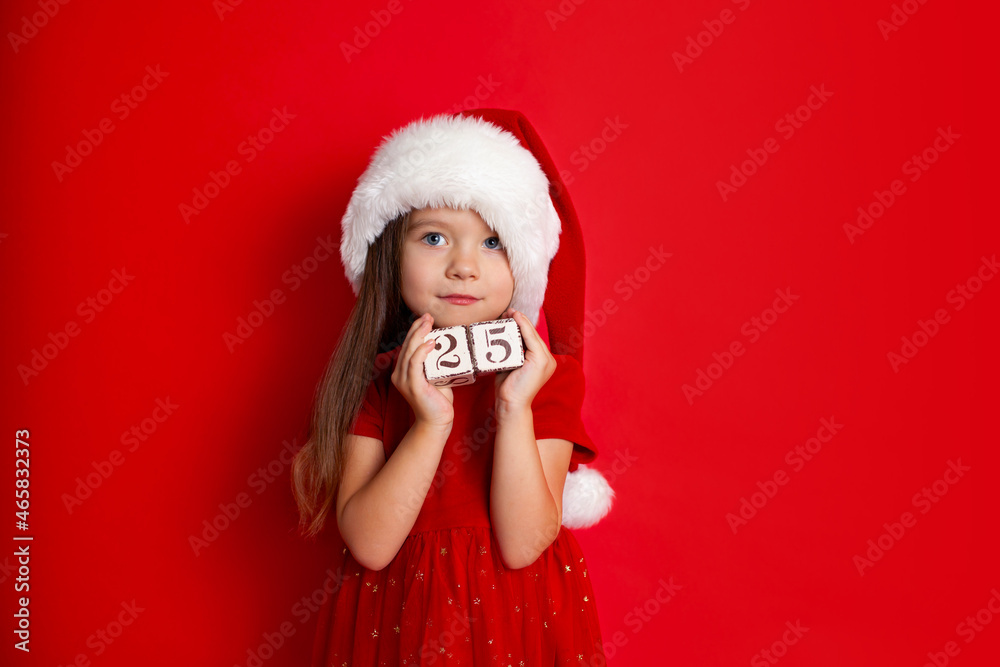 Merry Christmas and Happy Holidays. A funny girl in Santa's hat holds cubes with the numbers 25. Red background, space for text. Merry Christmas and Happy Holidays. A funny girl in Santa's hat holds cubes with the numbers 25. Red background, space for text.