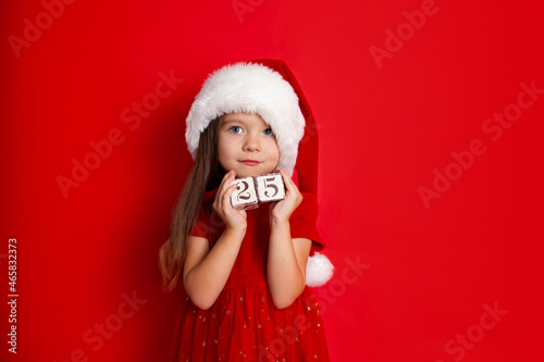 Merry Christmas and Happy Holidays. A funny girl in Santa s hat holds cubes with the numbers 25. Red background  space for text.