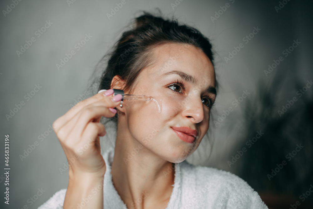 Brunette woman with long hair in a bun 30 years old applies serum on her face at home in the bathroom Brunette woman with long hair in a bun 30 years old applies serum on her face at home in the bathroom