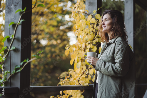  A young woman is drinking coffee and using the phone on the terrace in late autumn.