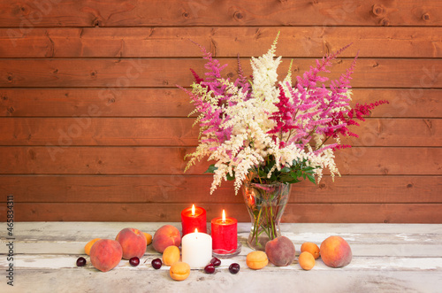 White, red and pink beautiful astilbe flowers in glass vase, burning candles and fresh fruits near  on aged wooden table.