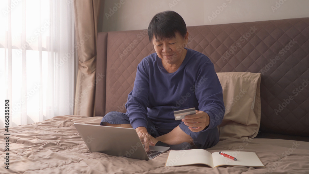 Happy elderly old Asian woman, people shopping online, using a credit card to pay money with a computer notebook laptop, business documents on bed in bedroom at home. Lifestyle. Happy elderly old Asian woman, people shopping online, using a credit card to pay money with a computer notebook laptop, business documents on bed in bedroom at home. Lifestyle.