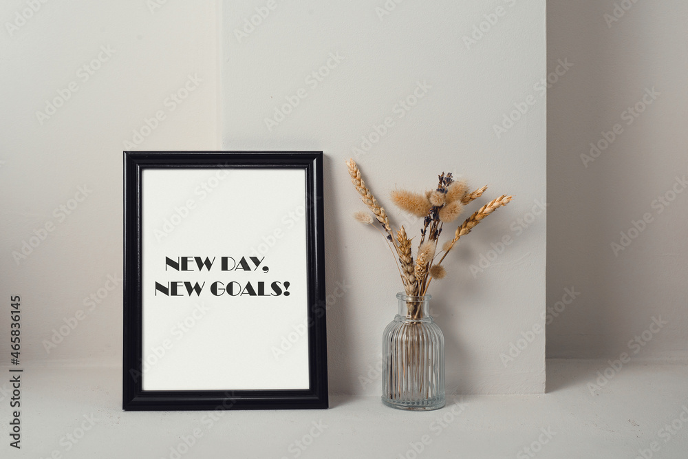 Motivational quote a new day, a new goal. Next to the black frame is a glass vase with dried herbs. Light background. Motivational quote a new day, a new goal. Next to the black frame is a glass vase with dried herbs. Light background.