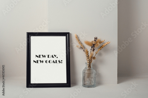 Motivational quote a new day, a new goal. Next to the black frame is a glass vase with dried herbs. Light background.