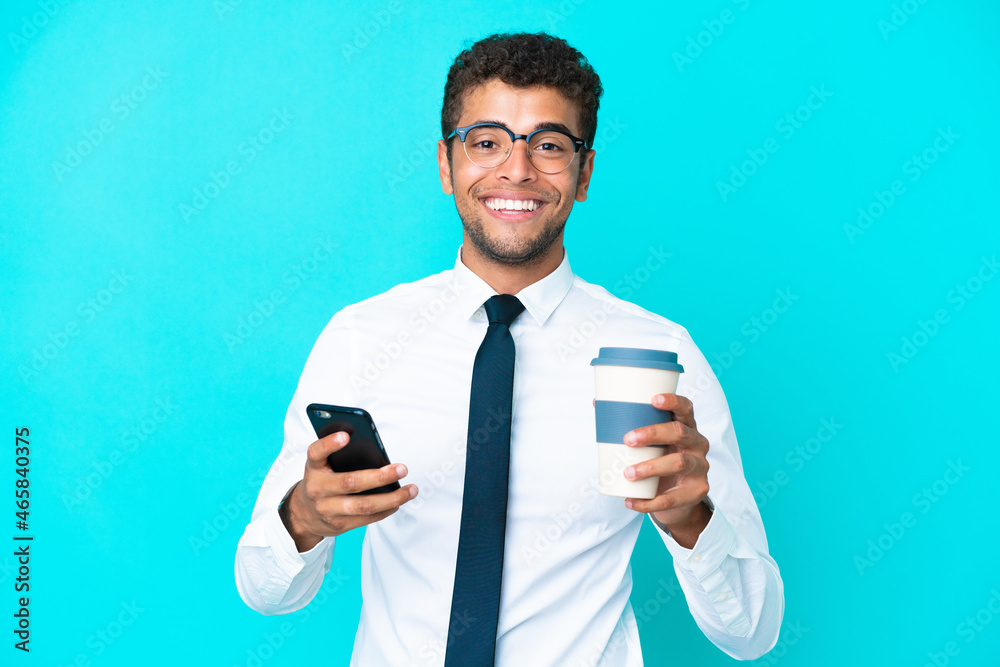 Young business Brazilian man isolated on blue background holding coffee to take away and a mobile Young business Brazilian man isolated on blue background holding coffee to take away and a mobile