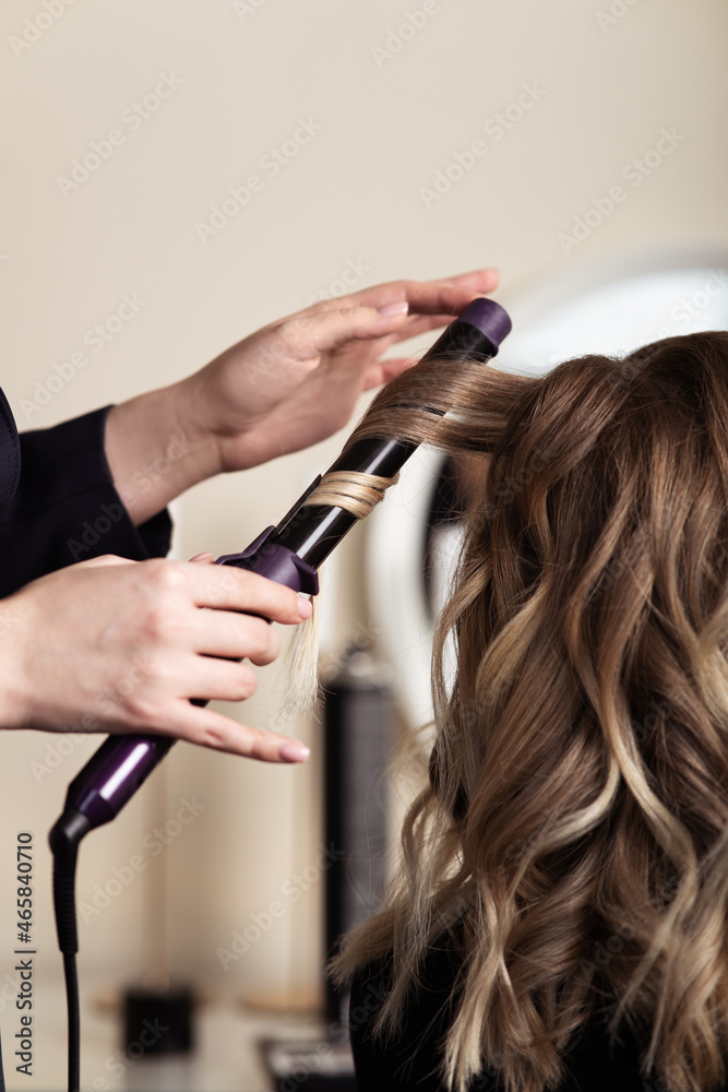 Fototapeta premium Female hands of a hair master. Close-up of blond hair on a curling iron. Curling curls.