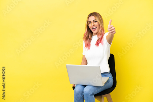 Young woman sitting on a chair with laptop over isolated yellow background with thumbs up because something good has happened