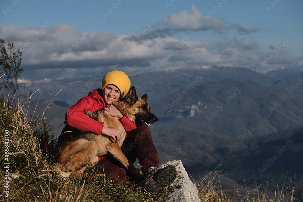Travel with German shepherd and owner. Hugging best friend in nature. Young Caucasian woman in red jacket is sitting on top of mountain next to her dog and enjoying scenery at sunset.