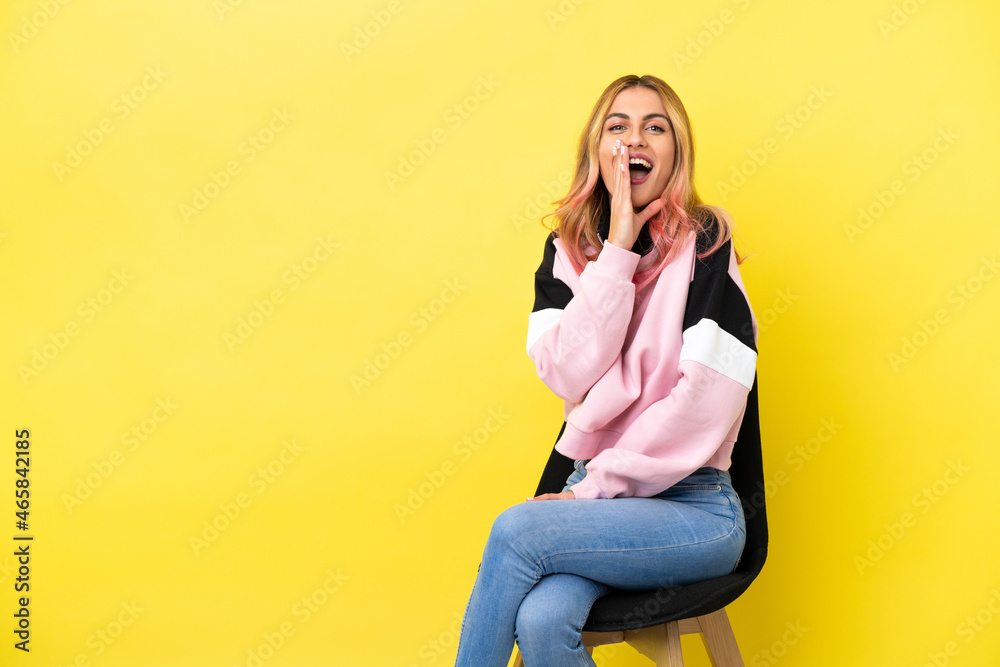 Young woman sitting on a chair over isolated yellow background shouting with mouth wide open Young woman sitting on a chair over isolated yellow background shouting with mouth wide open