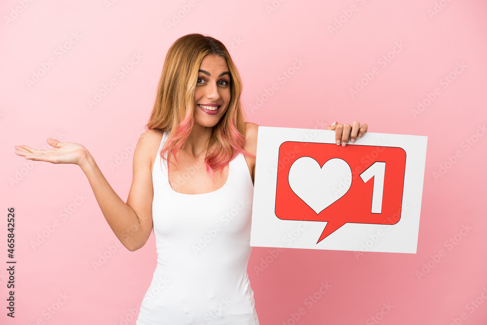 Young woman over isolated pink background holding a placard with Like icon with surprised expression