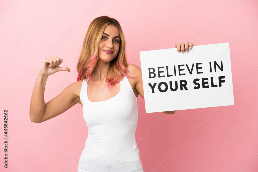 Young woman over isolated pink background holding a placard with text Believe In Your Self with proud gesture Young woman over isolated pink background holding a placard with text Believe In Your Self with proud gesture