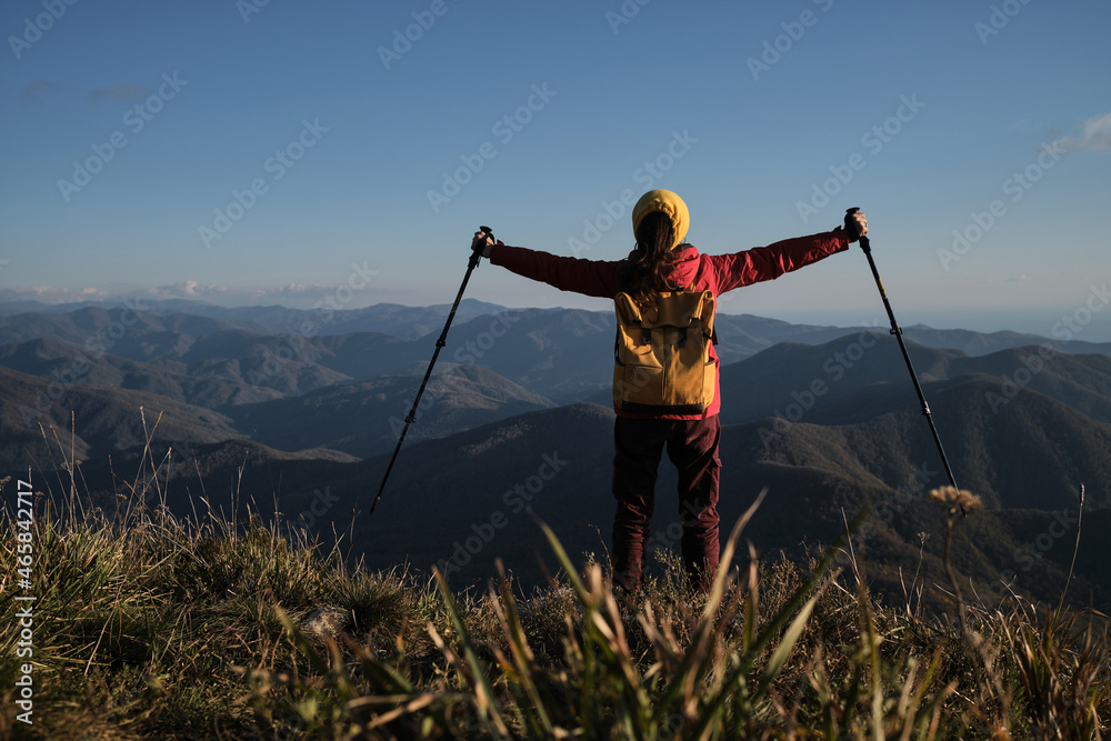 Travel in mountains alone. Beautiful panoramic view . Female traveler in red jacket with yellow backpack stands with back turned and looks at mountains with arms outstretched with sticks to sides.