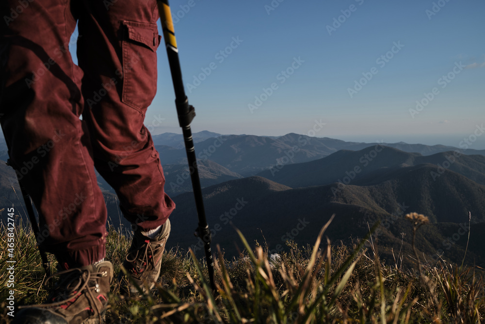 Trekking route in national park. Front view of feet in boots and hiking sticks. Main character is out of focus. Female traveler walks along narrow path on top of mountain among tall yellow dry grass. Trekking route in national park. Front view of feet in boots and hiking sticks. Main character is out of focus. Female traveler walks along narrow path on top of mountain among tall yellow dry grass.