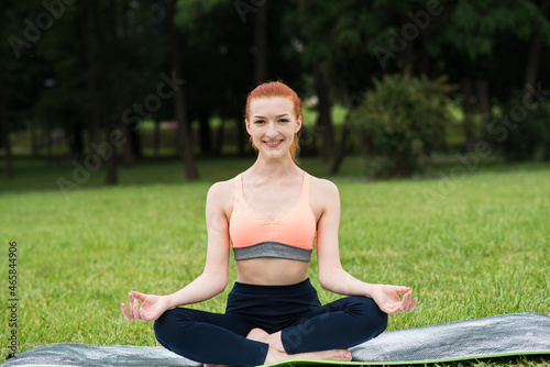 Redhead woman practicing yoga on a mat in the park on the grass. Sitting in lotus position