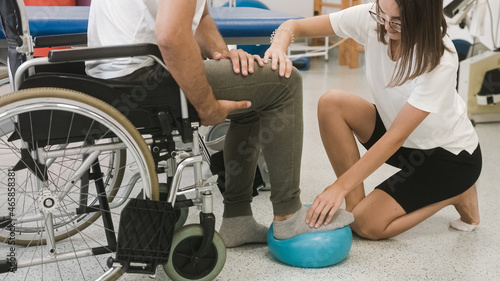 Fototapeta Naklejka Na Ścianę i Meble -  Pphysiotherapist and male patient seated in a wheelchair during rehabilitation treatment - press and release a foot on a small exercise ball