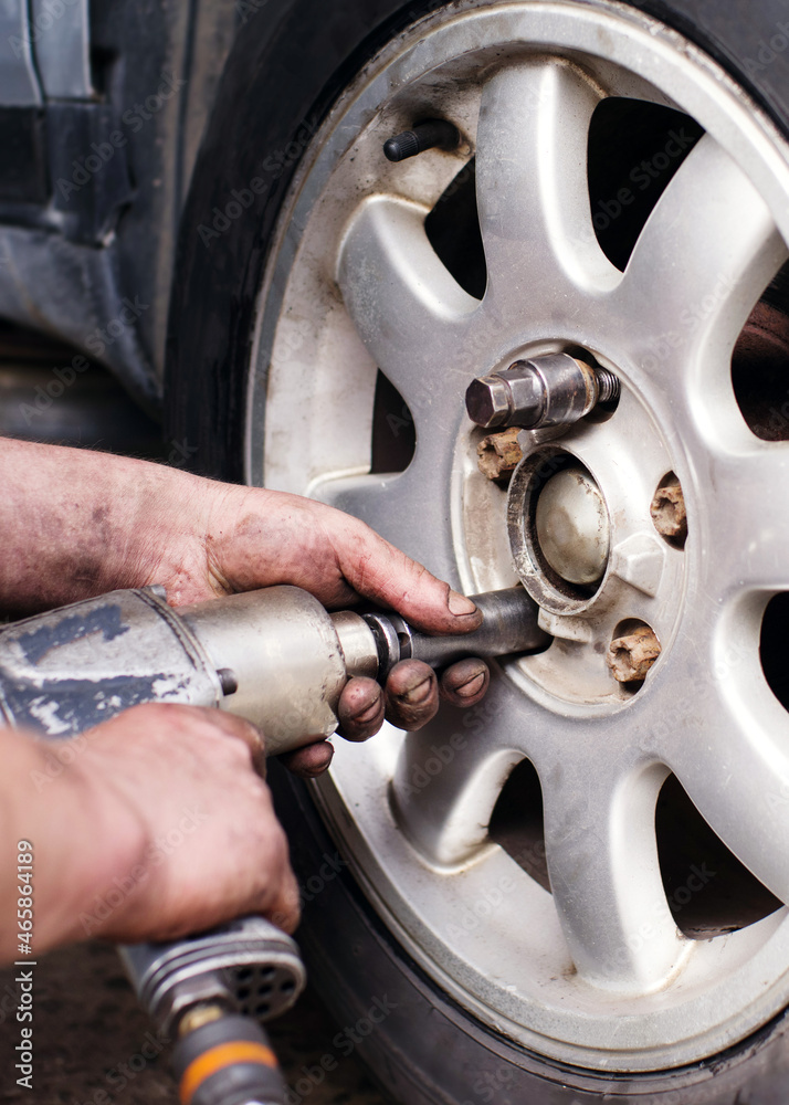 Fototapeta premium A mechanic at a service station replaces a car wheel, a worker's hands in black fuel oil and a tool for unscrewing bolts close-up near the wheel. Tire service, workflow