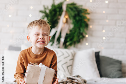 Cute happy Caucasian boy opening his presents on Christmas morning. Christmas tree wreath on background. Child excited and smiling. Happy family holidays at home. Opening gifts in boxes. Copy space.