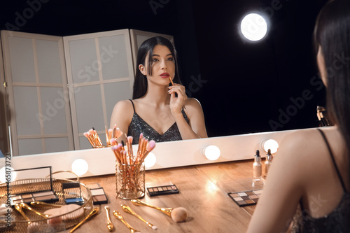 Beautiful young woman applying lipstick with brush in dressing room at night