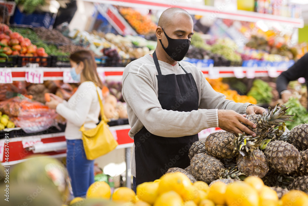 Latino-american worker in medical mask in supermarket with pineapples during COVID-19 pandemic