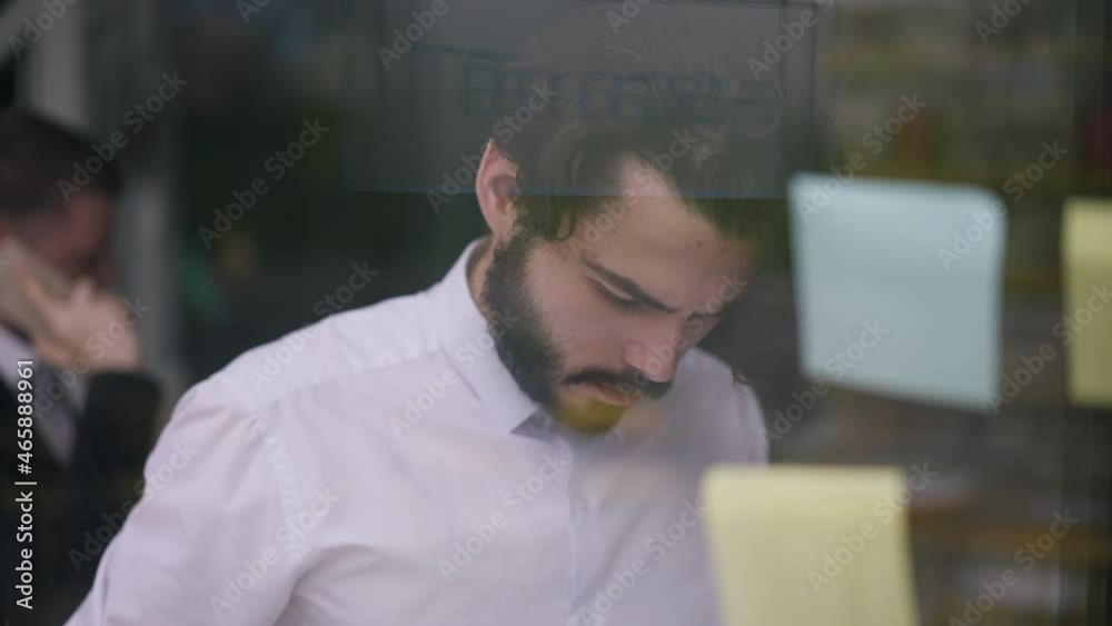 Concentrated bearded man writing ideas on adhesive notes on glass door thinking. Portrait of busy focused thoughtful Caucasian manager planning project. Slow motion