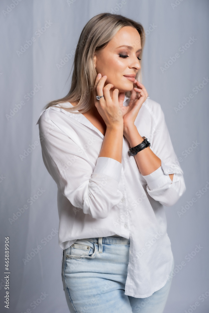 beautiful woman portrait in studio posing standing. girl in a white shirt and blue jeans, barefoot. blonde with straight hair and bright makeup. beauty concept. Gray background beautiful woman portrait in studio posing standing. girl in a white shirt and blue jeans, barefoot. blonde with straight hair and bright makeup. beauty concept. Gray background