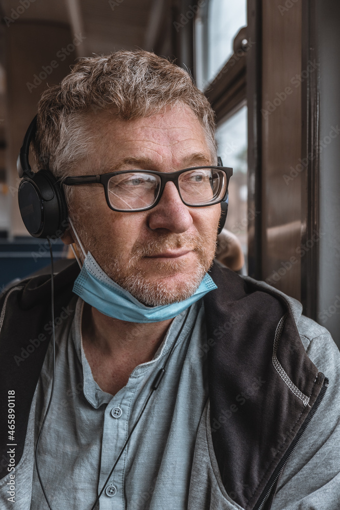 Portrait of an adult man wearing headphones and glasses with a medical protective mask on his chin on public transport. Improper use of a protective face mask. New normal concept portrait.