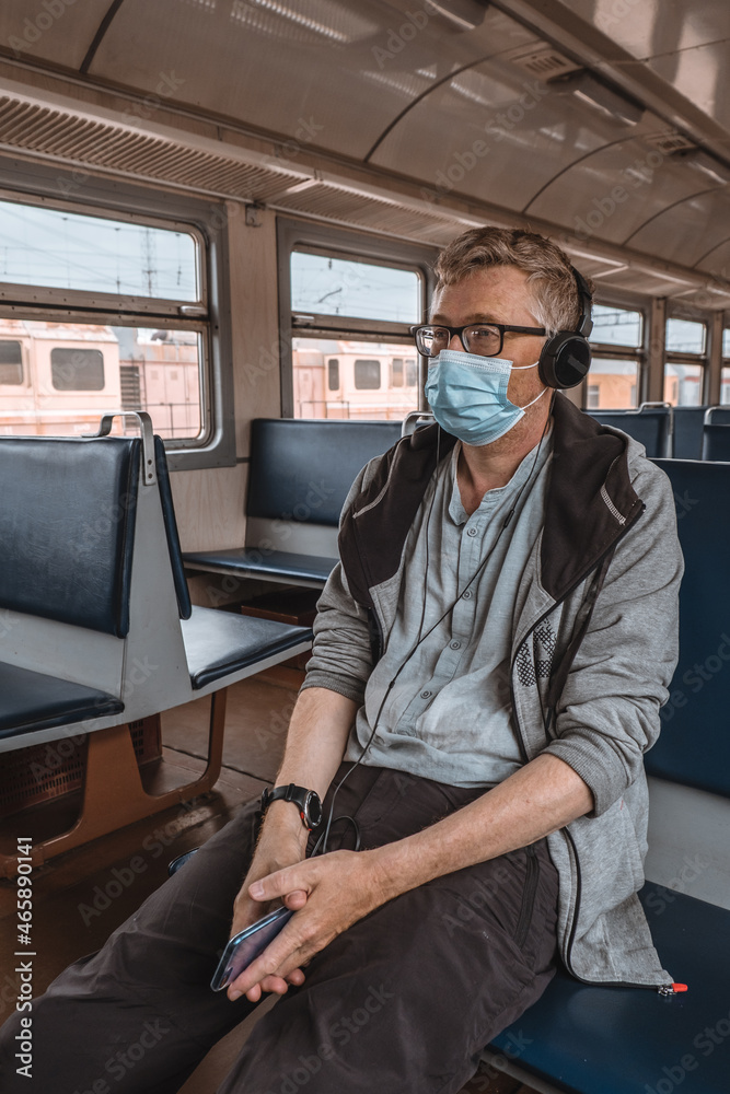 Portrait of a serious adult man in glasses and headphones with a medical protective mask on his face in an empty train car. Preventive measure. Social distance during the coronavirus pandemic.