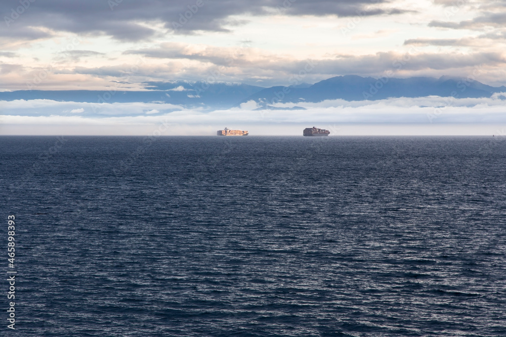 Distant Cargo Boats Nautical Vessels Sailing Strait of Juan De Fuca on Horizon. Coast of Victoria, BC on Vancouver Island with Distant Clouds over Olympic Peninsula, Washington USA