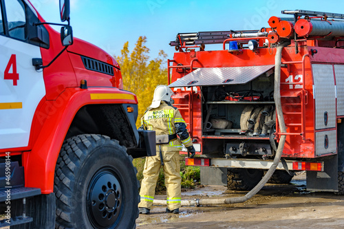 Rear view of a Russian firefighter in a protective suit and white helmet fighting a forest fire, a fire engine. Fire extinguishing operation. Behind a sign with the words 