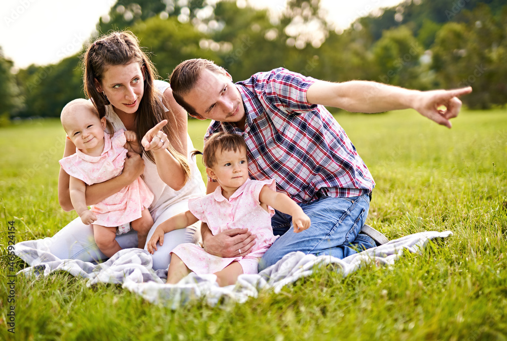 Happy family with young, millennial parents and two baby girls sitting on a picnic blanket and  pointing on something in the distance to show it to their children - Outdoor lifestyle family concept