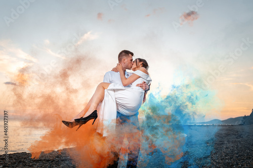 Wedding. Groom holds the bride in his arms and kisses her, surrounded by colorful smoke. There is an ocean and a sunset in the background. Valentine's Day