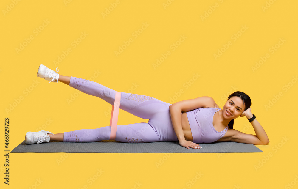 Full length of african american woman doing exercises with elastic band, training her leg muscles over yellow background Full length of african american woman doing exercises with elastic band, training her leg muscles over yellow background