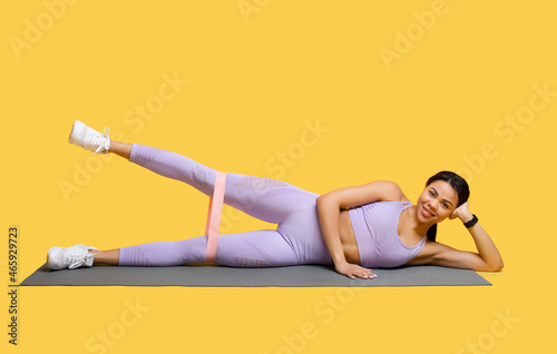 Full length of african american woman doing exercises with elastic band, training her leg muscles over yellow background