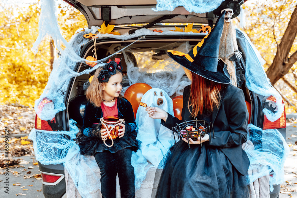 siblings teenage girl in witch costume and hat, cute little girl in spooky costume and cute poodle dog in ghost costume sits in trunk car decorated for Halloween with web, orange balloons and pumpkin