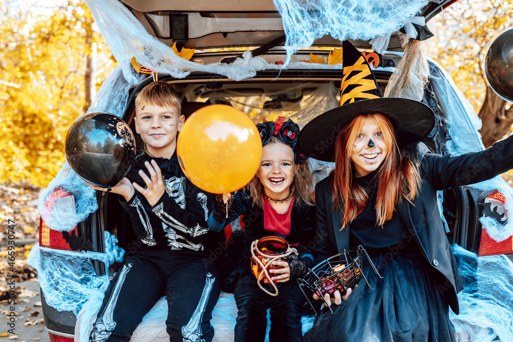 siblings boy in skeleton costume, teenage girl in witch costume and hat and cute little girl in spooky costume sits in trunk car decorated for Halloween with web, orange balloons and pumpkins, outdoor - obrazy, fototapety, plakaty 