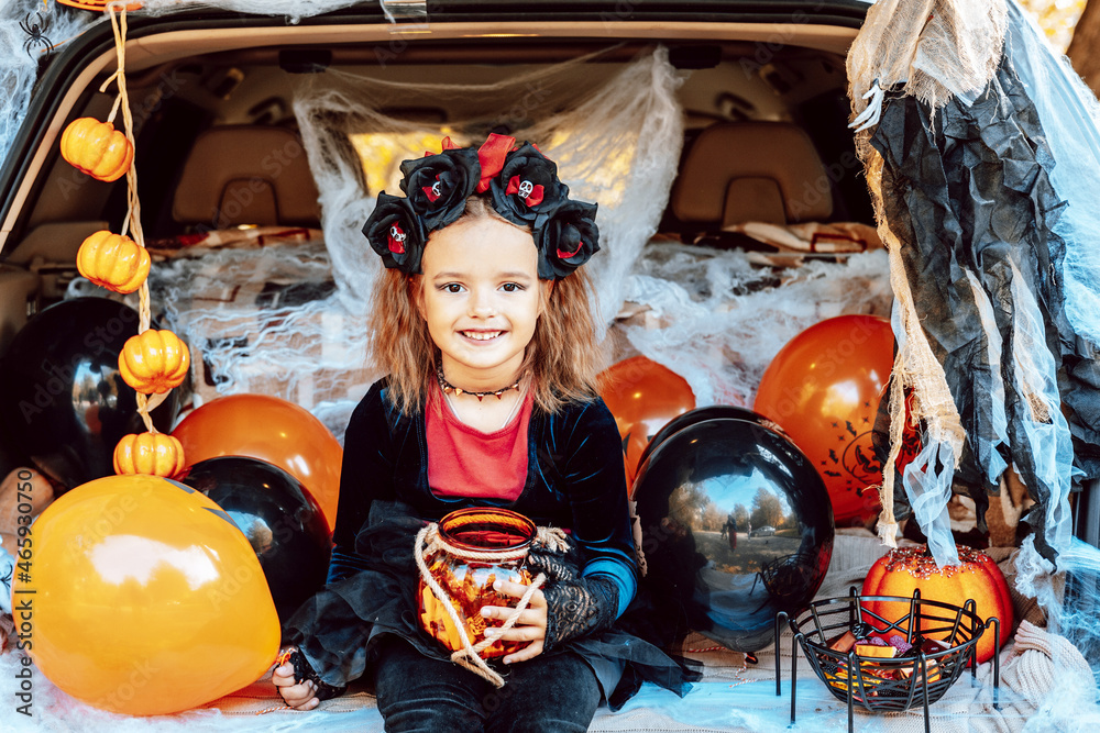 little girl in spooky costume and hat with bucket of sweets and cute poodle dog in ghost costume sits in trunk car decorated for Halloween with web, orange balloons and pumpkins, outdoor creative