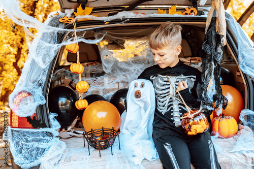 preteen caucasian boy in skeleton costume with bucket of sweets and cute poodle dog in ghost costume sits in trunk car decorated for Halloween with web, orange balloons and pumpkins, outdoor creative