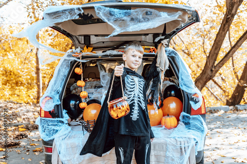 preteen cute caucasian boy in skeleton costume with bucket of sweets sits in trunk car decorated for Halloween with web, orange balloons and pumpkins, outdoor creative