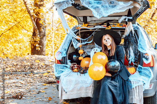 teenage girl in witch costume and hat and cute poodle dog in ghost costume sits in trunk car decorated for Halloween with web, orange balloons and pumpkins, outdoor creative activity concept in autumn