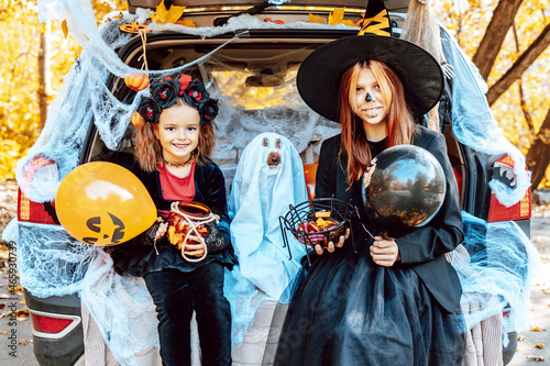 siblings teenage girl in witch costume and hat, cute little girl in spooky costume and cute poodle dog in ghost costume sits in trunk car decorated for Halloween with web, orange balloons and pumpkins © klavdiyav