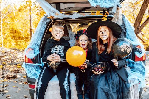 siblings boy in skeleton costume, teenage girl in witch costume and hat and cute little girl in spooky costume sits in trunk car decorated for Halloween with web, orange balloons and pumpkins, outdoor