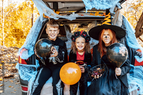 siblings boy in skeleton costume, teenage girl in witch costume and hat and cute little girl in spooky costume sits in trunk car decorated for Halloween with web, orange balloons and pumpkins, outdoor