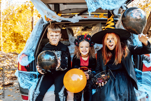 siblings boy in skeleton costume, teenage girl in witch costume and hat and cute little girl in spooky costume sits in trunk car decorated for Halloween with web, orange balloons and pumpkins, outdoor