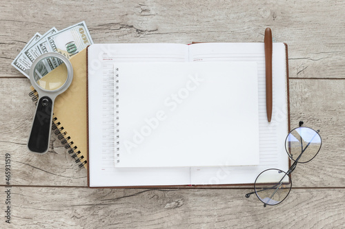 Flat lay of pencil notepad with place for text. magnifier and notepad with money bills with blank center on wooden background, top view