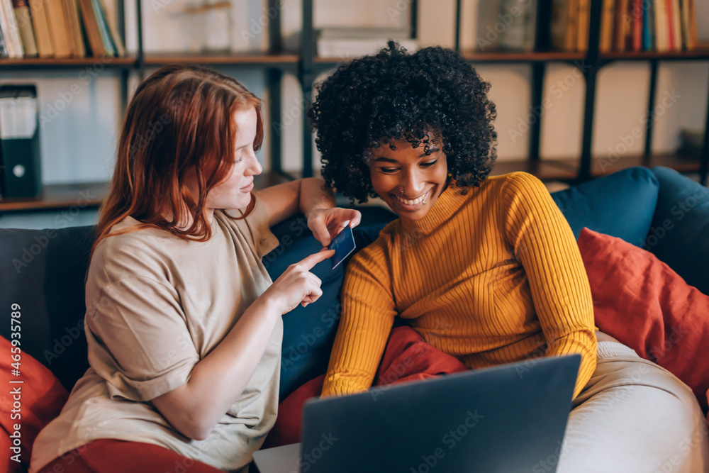 two cheerful young women friends of different ethnicity using discount card and laptop - young people surfing the net - making payments or shopping online two cheerful young women friends of different ethnicity using discount card and laptop - young people surfing the net - making payments or shopping online