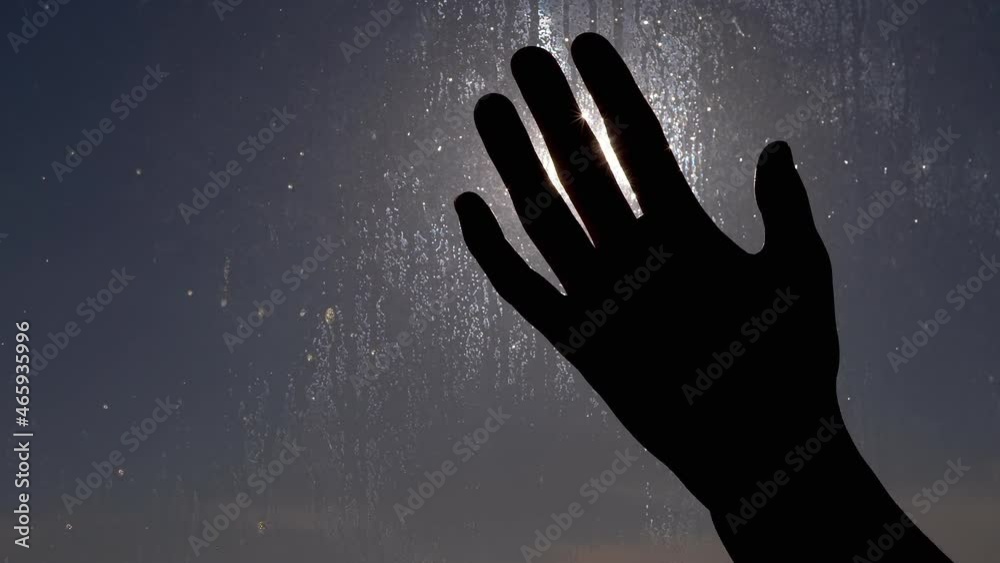Female Hand Plays with Rays of Sunlight on a Background of Wet Glass. Fingers touching bright light of sunlight through a closed window at sunset. Raindrops run down the glass in the glare of the sun.